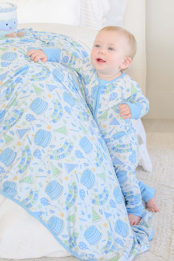 A baby wearing a Caden Lane light blue and white footed sleeper with a colorful birthday-themed pattern, sitting on a white bed and holding a Caden Lane matching birthday patterned blanket.