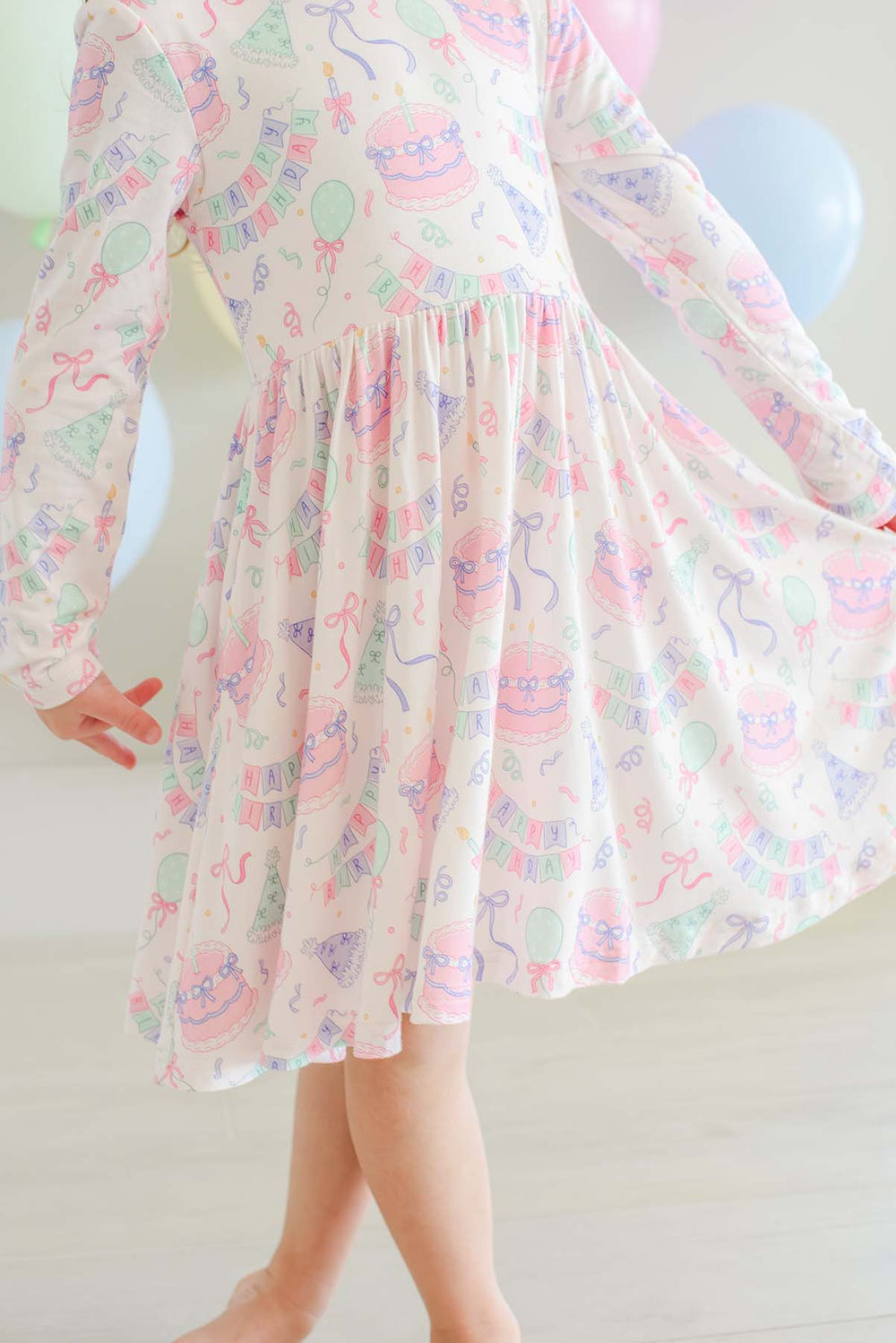 A child wearing a Caden Lane long-sleeve white dress with a pastel multicolor print of cakes, balloons, and party decorations, shown from the shoulders down in a bright indoor setting.