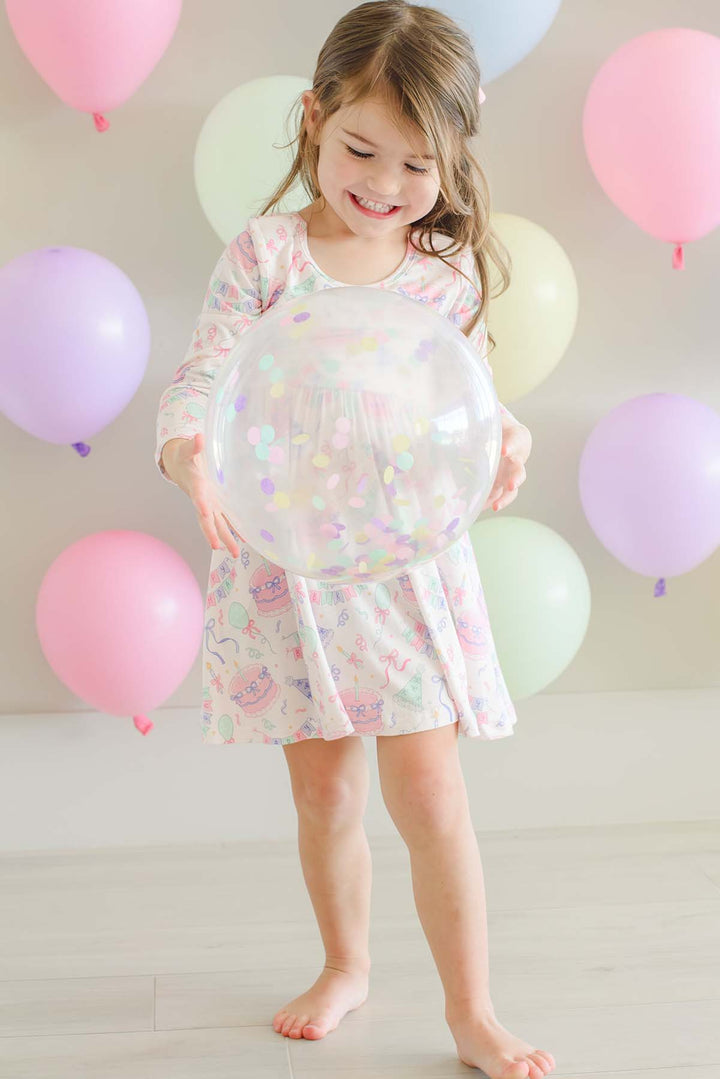 A young girl with light brown hair stands barefoot on a light wood floor, holding a large clear inflatable ball decorated with pastel confetti. She is wearing a Caden Lane long-sleeve, knee-length white dress with a colorful pastel unicorn and cat pattern.