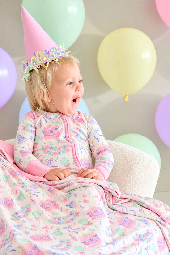 Smiling toddler wearing Caden Lane Birthday Bash Pink bamboo pajamas and wrapped in a matching blanket with birthday cakes, balloons and party hats.