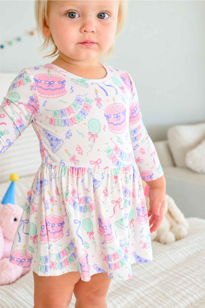 A toddler girl wearing a Caden Lane long-sleeve white dress with a colorful pastel birthday print, standing on a bed in a softly lit room.