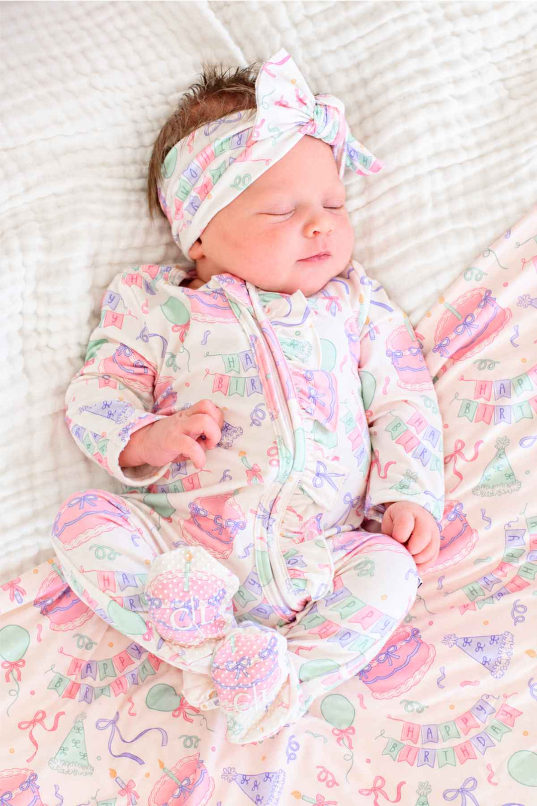 A sleeping newborn baby wearing a Caden Lane pastel multicolor party-themed footed sleeper and matching bow headband, lying on a white textured blanket.