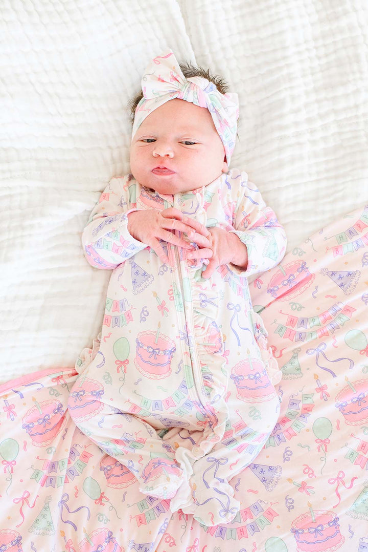 A newborn baby lying on a white textured blanket, wearing a Caden Lane light pink footed sleeper with a matching bow headband and a matching blanket printed with pastel birthday-themed illustrations.