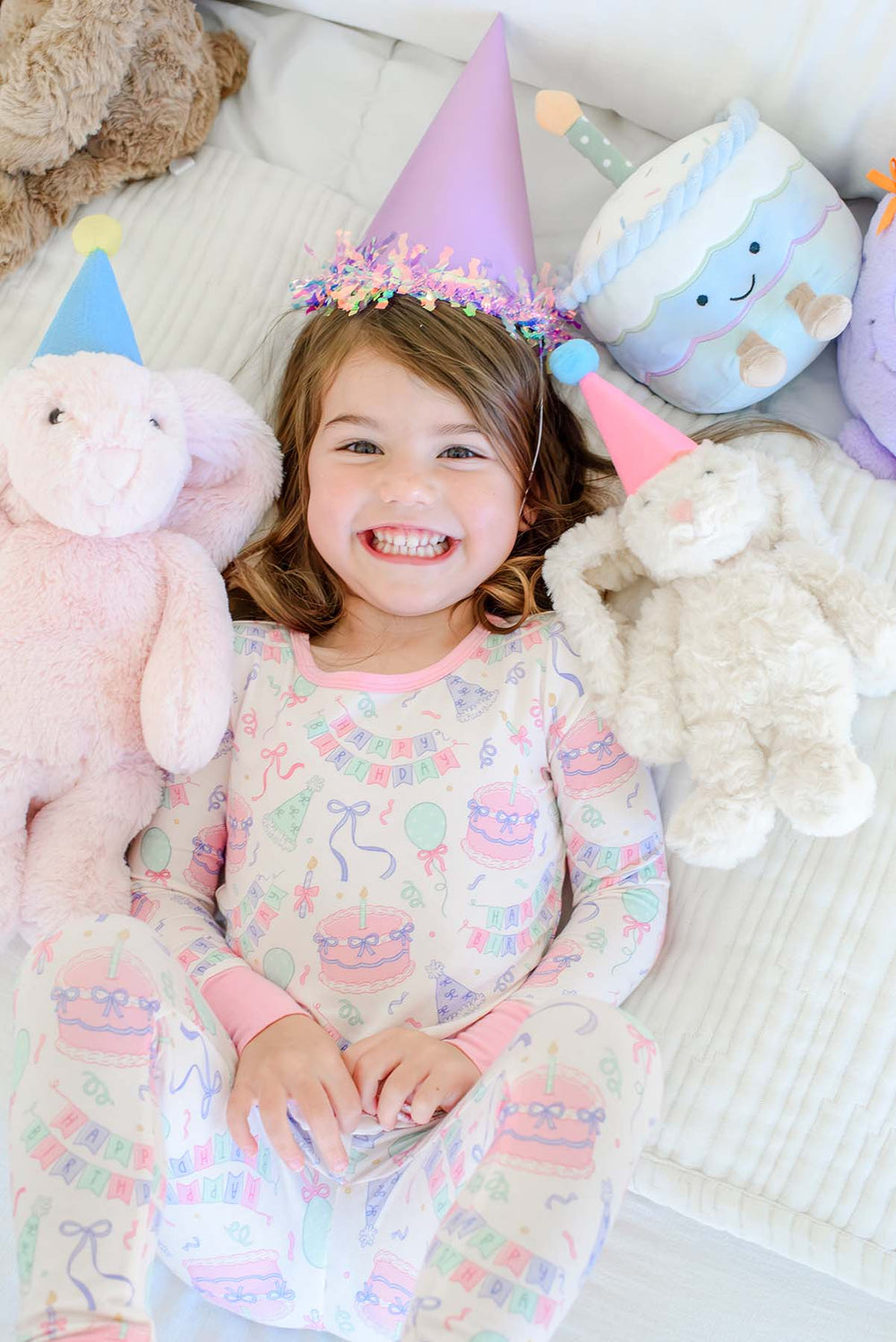 Overhead view of a smiling little girl wearing a Caden Lane Birthday Bash two-piece pajama set in pink with cake and bunting print, lying between plush bunny toys and a plush birthday cake toy, all wearing tiny party hats.