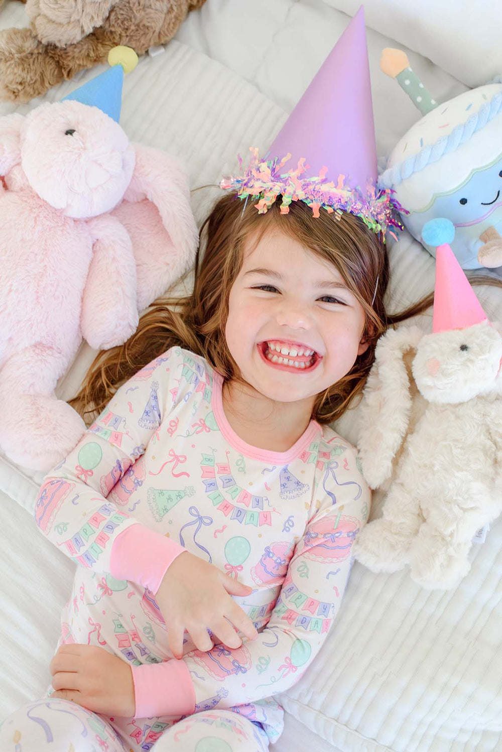 A young girl smiling while wearing a Caden Lane multicolor birthday pajama set and a party hat, lying on a white bed surrounded by plush stuffed animals.