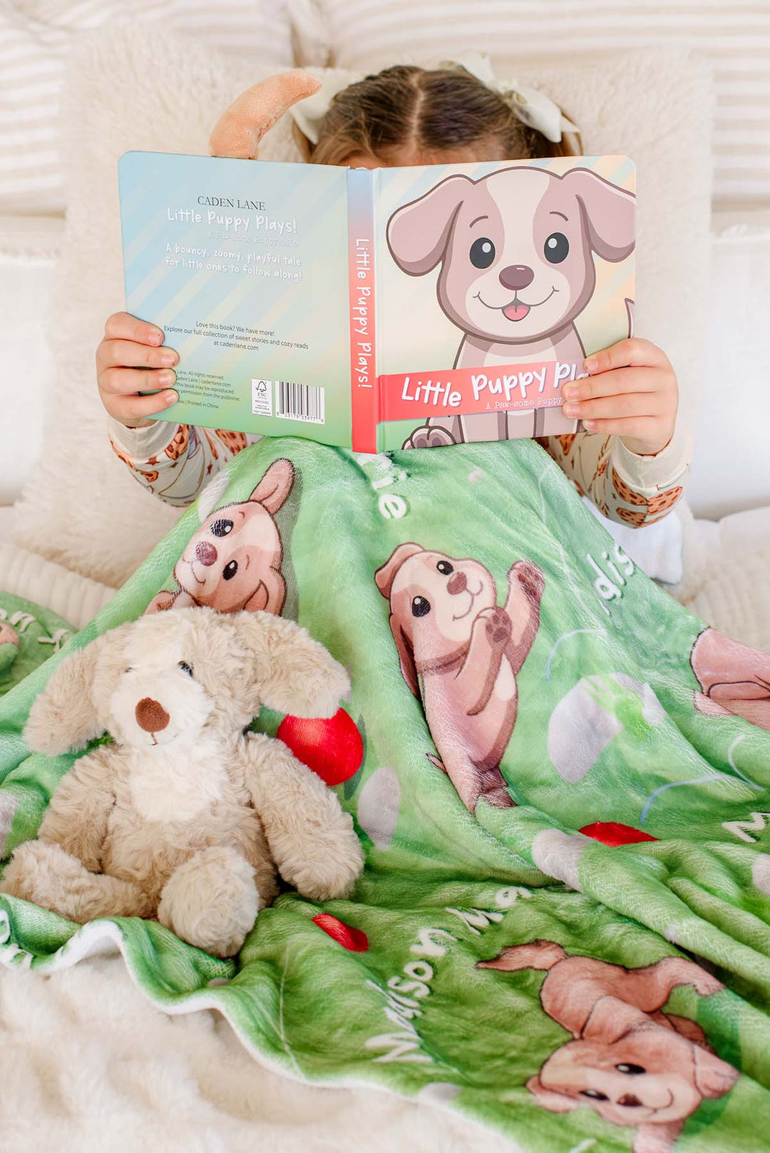Little girl holding up the open "Little Puppy Plays!" board book to cover her face, sitting with a plush puppy toy on a Caden Lane personalized green puppy-print blanket