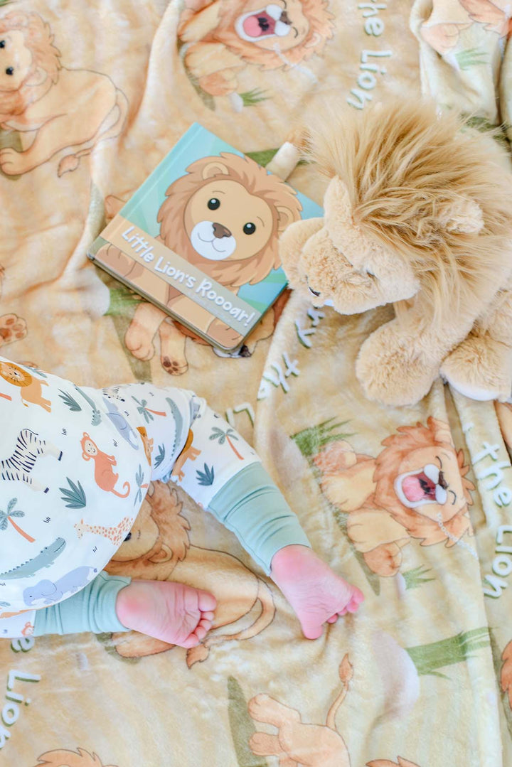 Close-up of a baby's feet on a Caden Lane personalized "Rory the Lion" fleece blanket, with a plush lion toy and the "Little Lion's Roooar!" board book nearby