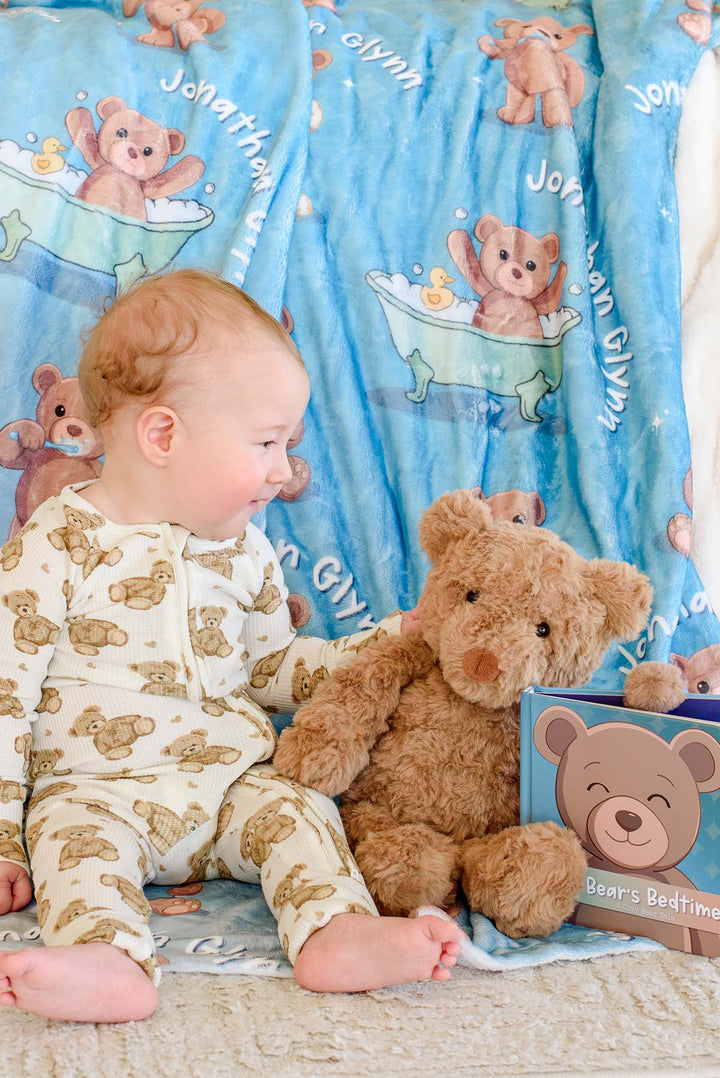 Baby boy in teddy bear pajamas sitting and reaching for a plush teddy bear in front of the "Little Bear's Bedtime" board book, with a Caden Lane personalized blue bear-print blanket in the background.