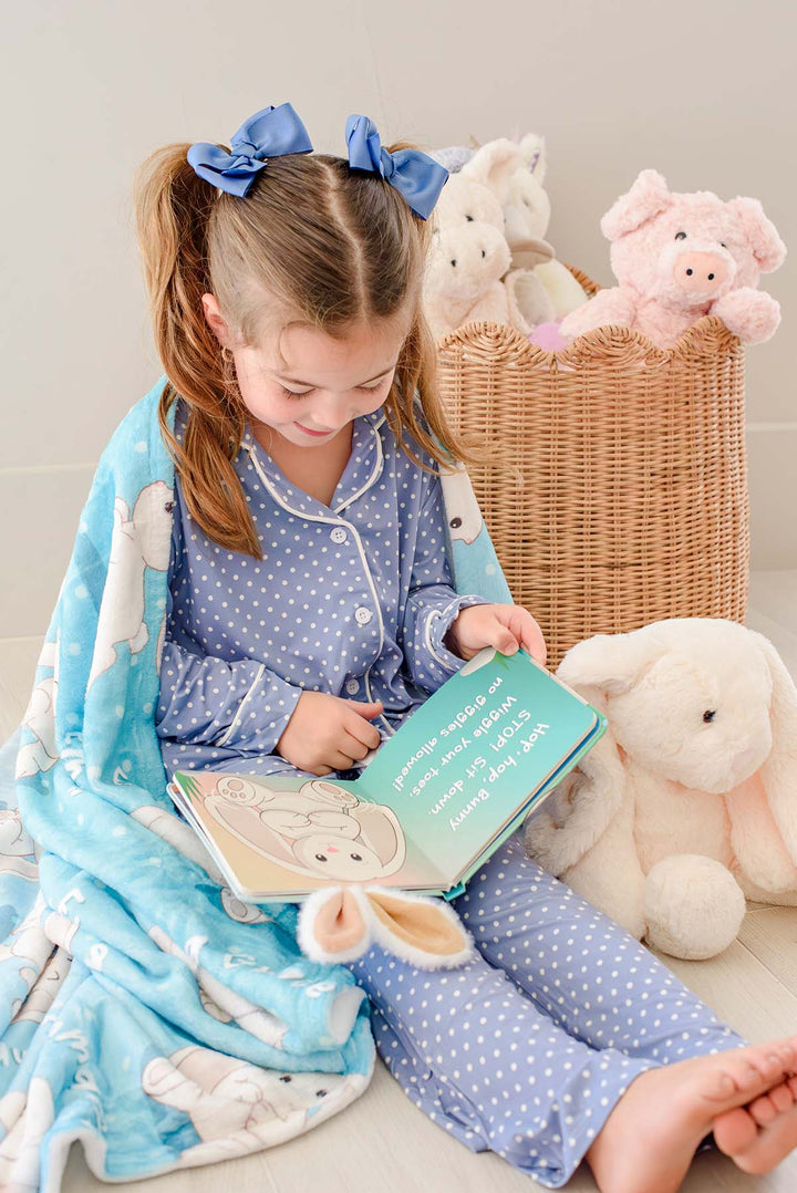 Little girl in blue polka-dot pajamas sitting and reading the open "Hop! Hop! Bunny!" board book, wrapped in a Caden Lane personalized blue bunny-print blanket with a plush bunny and basket of stuffed animals nearby.