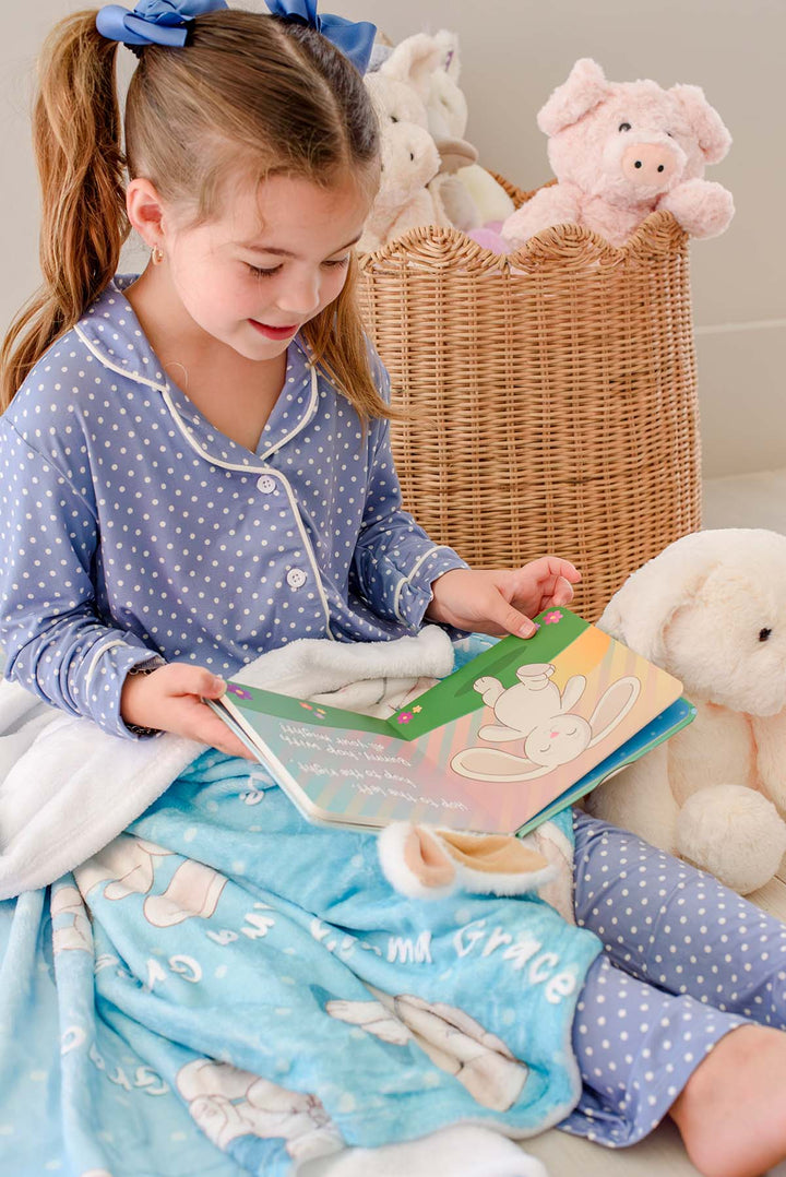 Little girl in blue polka-dot pajamas reading the open "Hop! Hop! Bunny!" board book, sitting on a Caden Lane personalized blue bunny-print blanket with a plush bunny and basket of stuffed animals nearby.