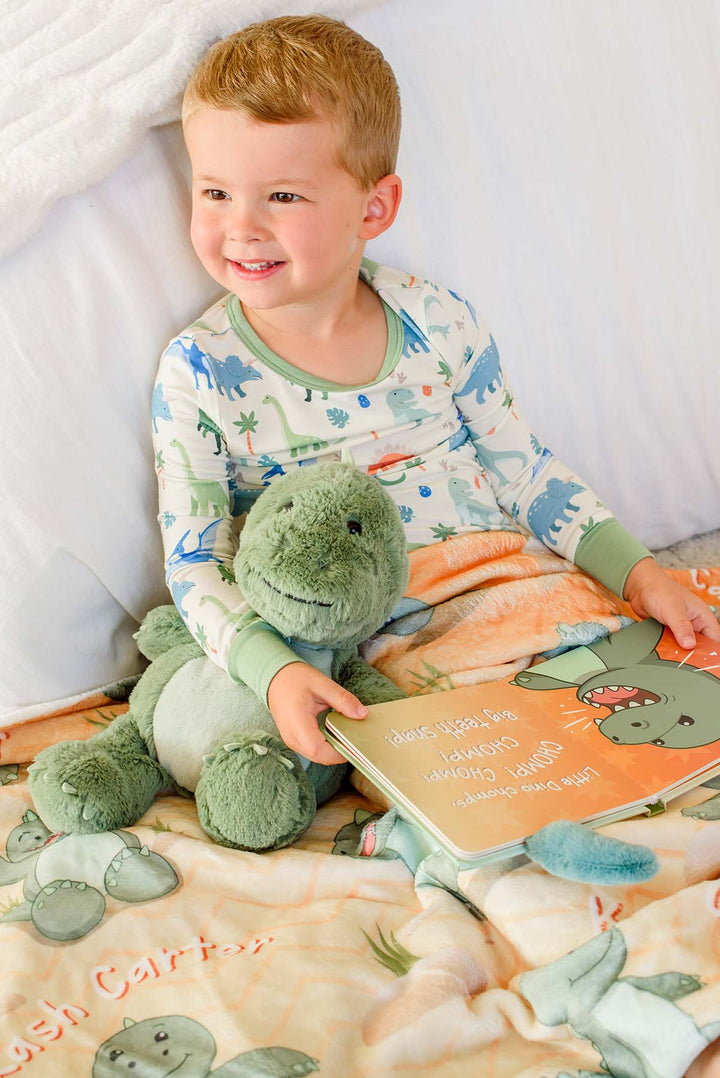 Toddler boy in Caden Lane dinosaur pajamas sitting up in bed smiling, holding a plush green dinosaur and an open "Little Dino Stomps!" board book, resting on a Caden Lane personalized orange dinosaur-print blanket.