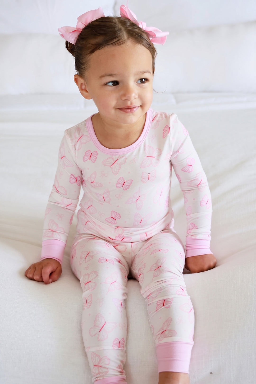 Two Piece Pajama Set in Blooming Butterflies print - smiling toddler sitting on white surface wearing the pink butterfly print long-sleeve top and pants set with pink bow headband