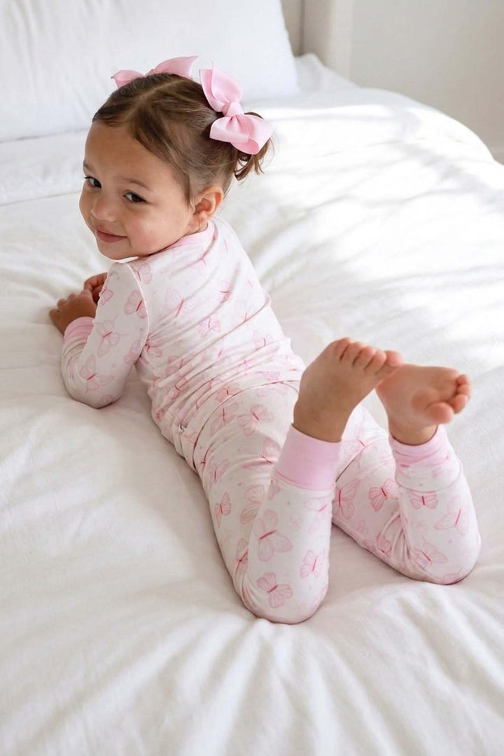 Two Piece Pajama Set in Blooming Butterflies print - toddler lying playfully on a white bed wearing the butterfly pajama set, with pink bow headband and cuffed ankles visible