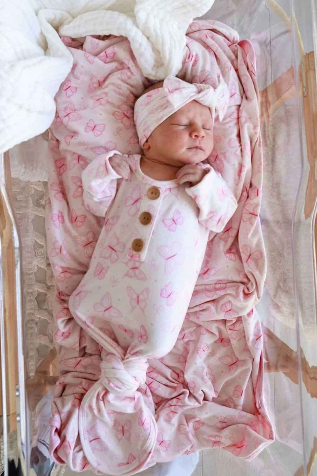 Newborn Baby Knot Gown & Hat Set in Blooming Butterflies print - sleeping newborn wearing the pink butterfly-print knot gown with gold snaps and matching knotted hat in a bassinet