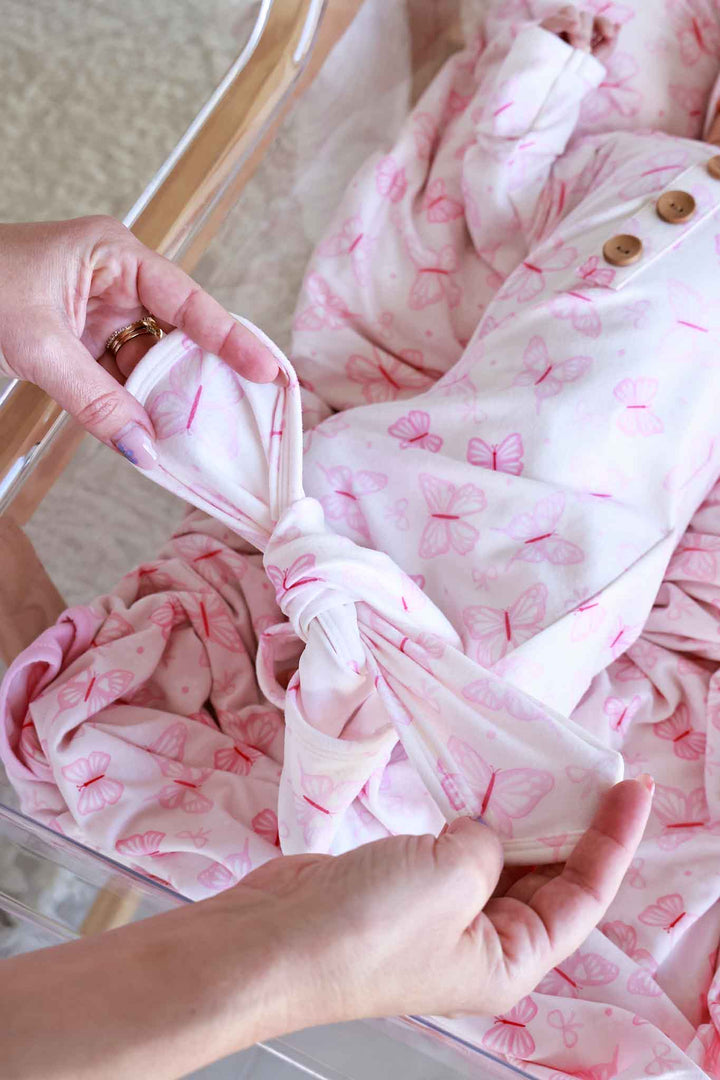 Newborn Baby Knot Gown & Hat Set in Blooming Butterflies print - caregiver tying the gown's knot bottom on baby in bassinet, demonstrating the easy-tie leg opening feature