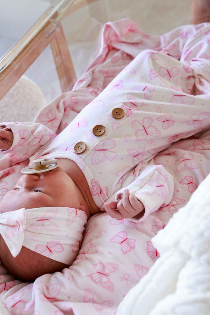Newborn Baby Knot Gown & Hat Set in Blooming Butterflies print - close-up of newborn wearing the gown and matching hat in bassinet, with gold button snaps and butterfly print visible