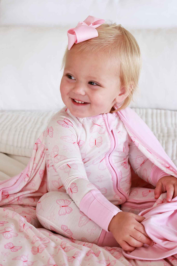 Convertible Zip Romper in Blooming Butterflies print - smiling toddler sitting on a couch wearing the pink butterfly zip romper, with pink bow hair accessory