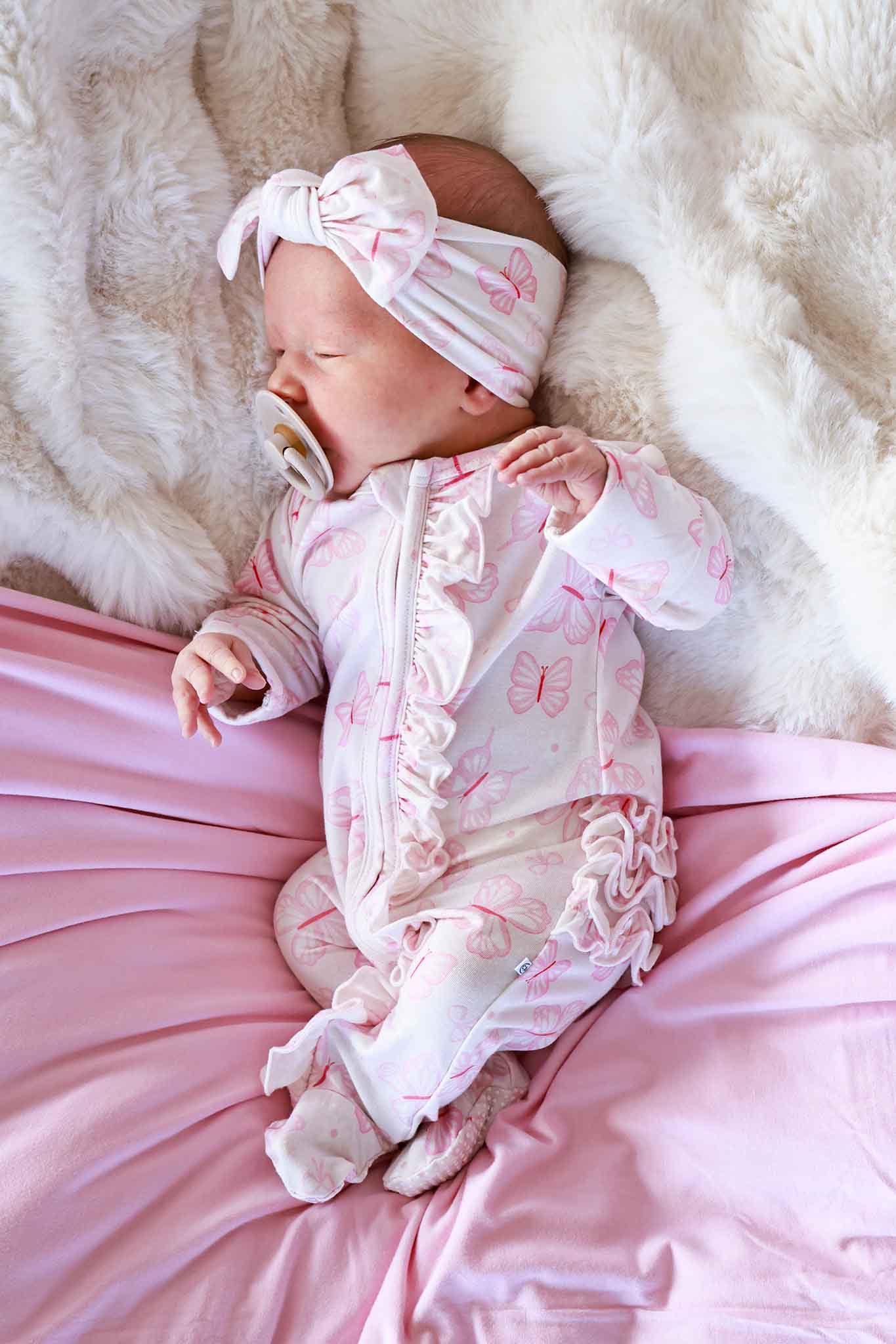 Ruffle Zipper Footie in Blooming Butterflies print - newborn lying on a white fluffy surface wearing the pink butterfly-print footie with ruffle trim and matching bow headband