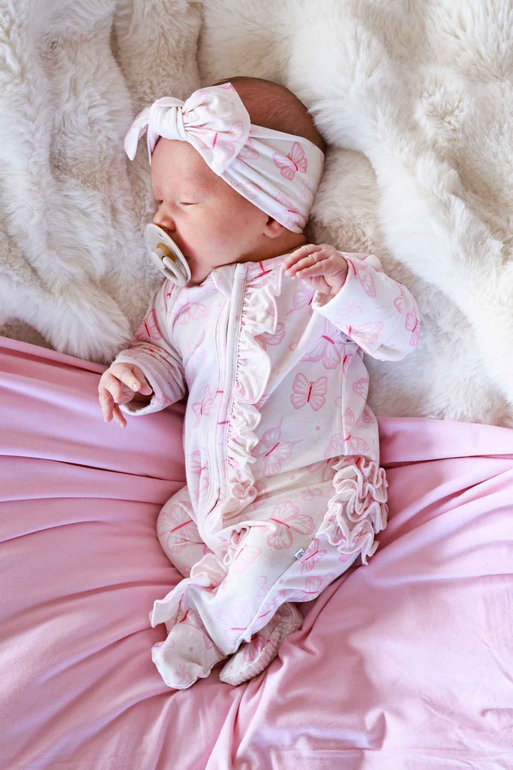 Ruffle Zipper Footie in Blooming Butterflies print - newborn lying on a white fluffy surface wearing the pink butterfly-print footie with ruffle trim and matching bow headband