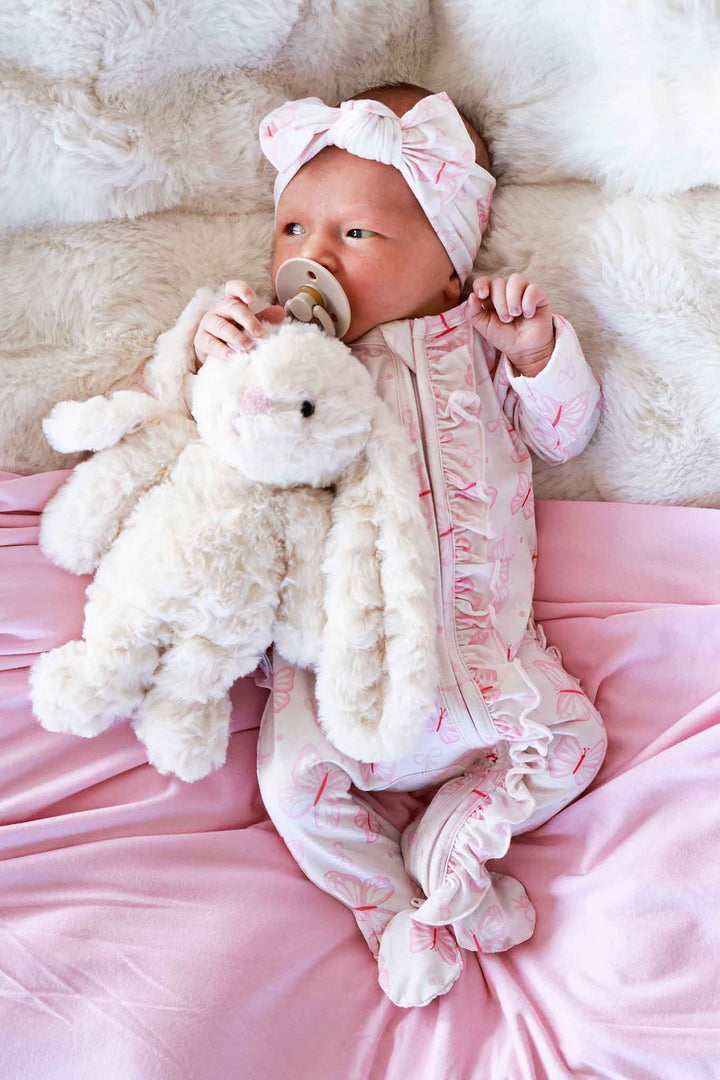 Ruffle Zipper Footie in Blooming Butterflies print - newborn lying on pink satin holding a stuffed bunny, wearing the butterfly print footie with bow headband and ruffle trim visible