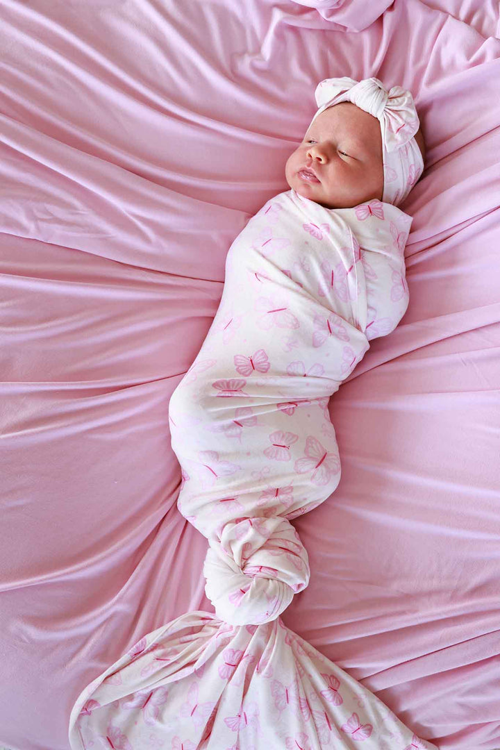 Oversized Swaddle Blanket in Blooming Butterflies print - newborn loosely wrapped in the light pink butterfly-print oversized swaddle on satin, with a white bow headband