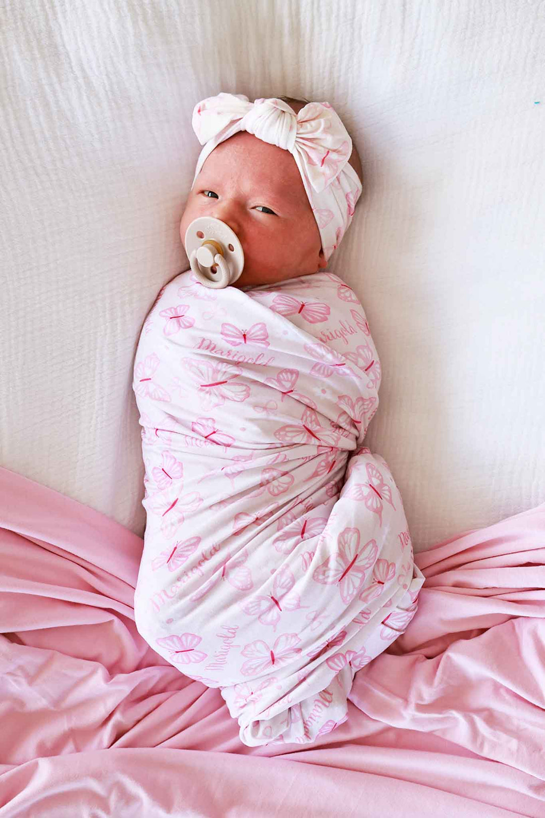 Personalized Swaddle Blanket in Blooming Butterflies print - newborn tightly swaddled in the pink print blanket on satin pink sheets, with a matching bow turban headband