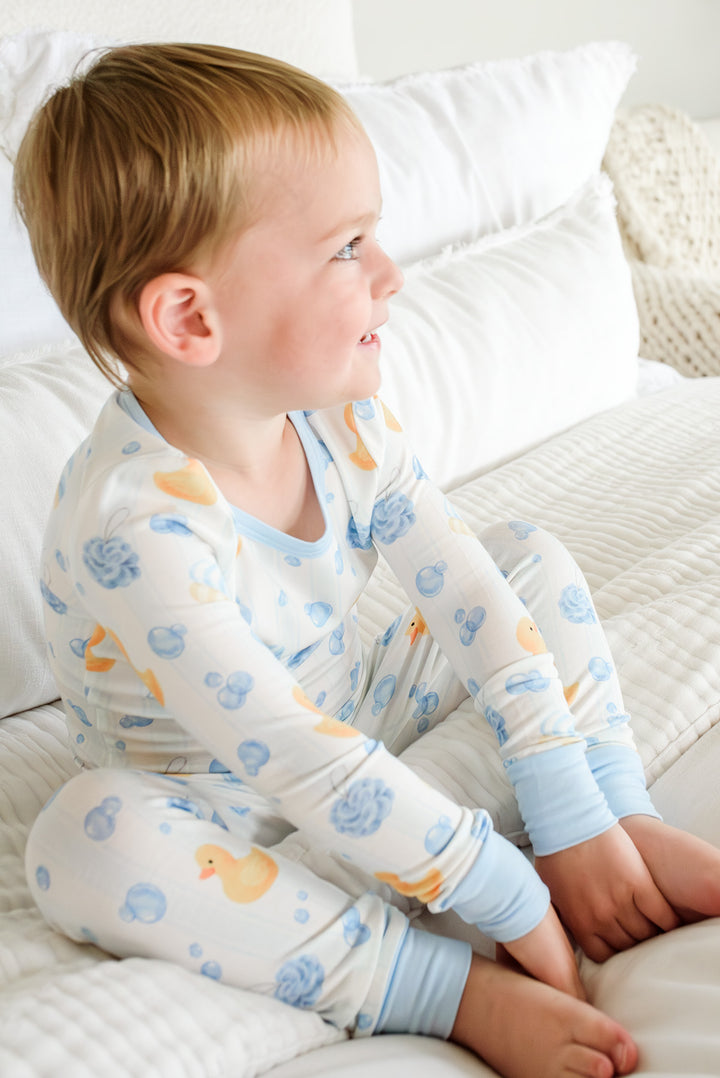 Boy sitting on bed looking sideways in blue Bubble Duckies two-piece bamboo pajamas with yellow rubber duck print