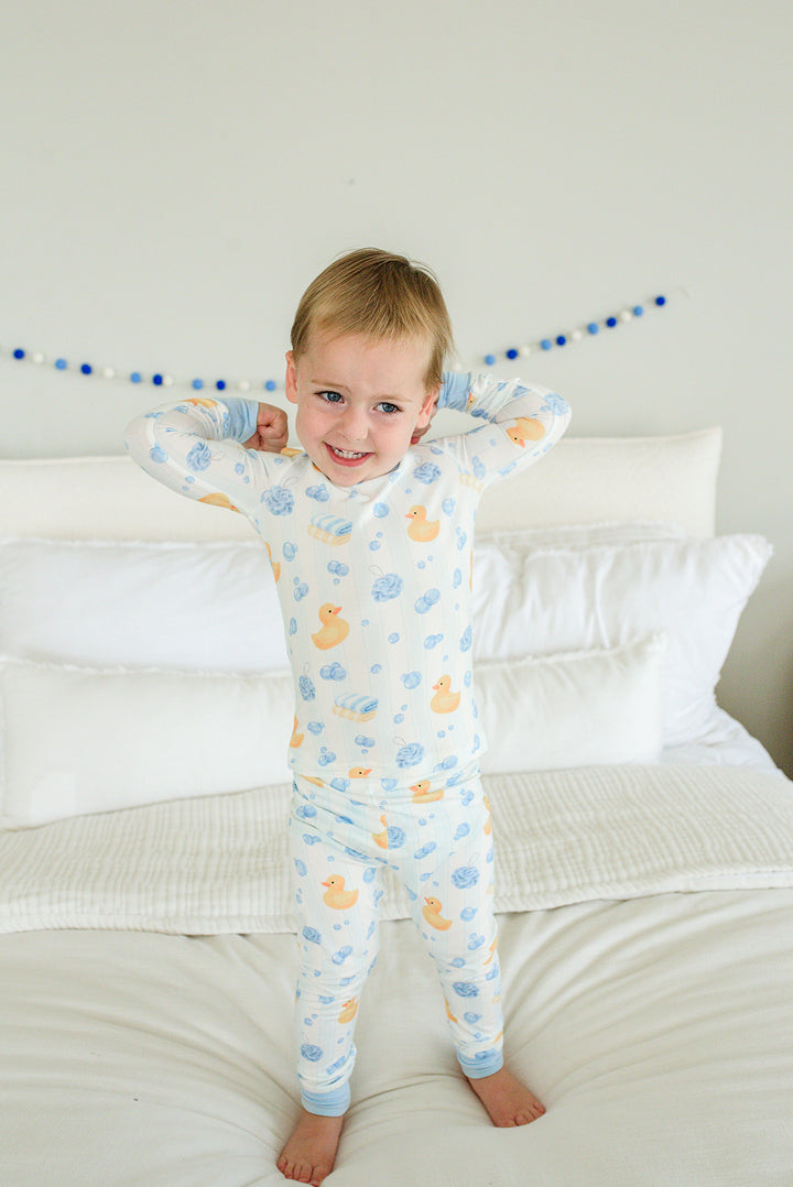 Toddler boy standing on bed with arms raised wearing blue Bubble Duckies two-piece bamboo pajamas