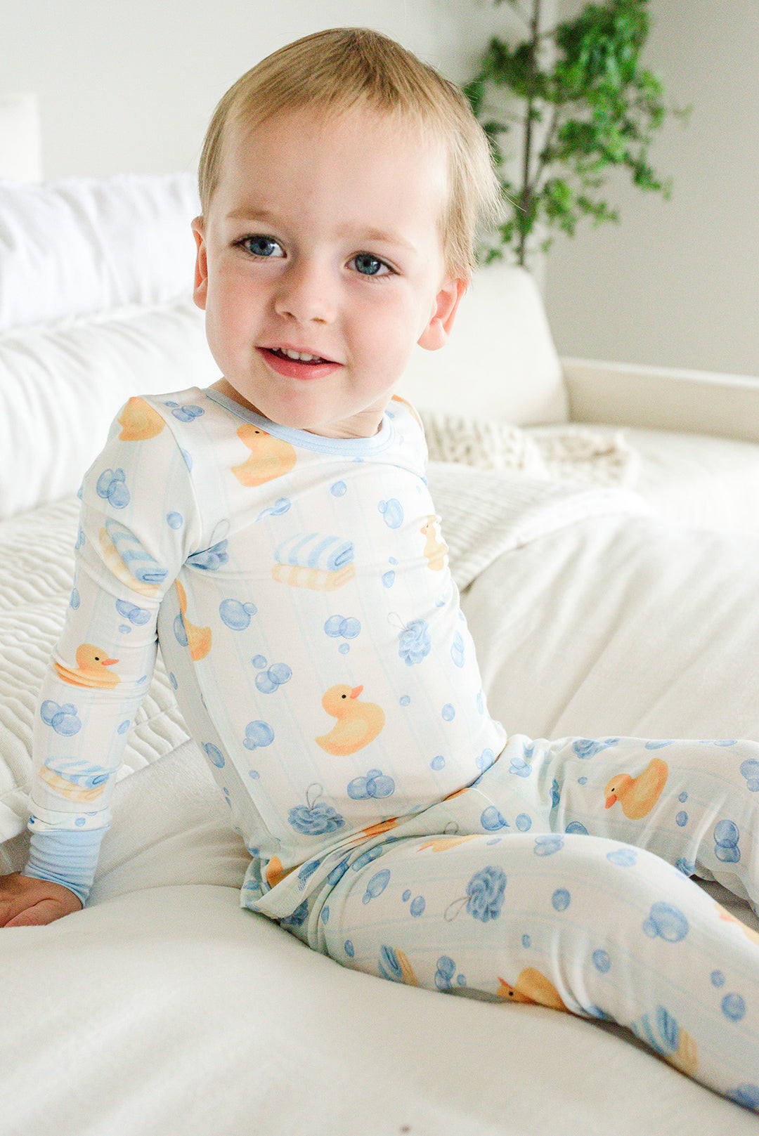 Young boy sitting on bed wearing blue Bubble Duckies two-piece bamboo pajamas with yellow rubber duck print