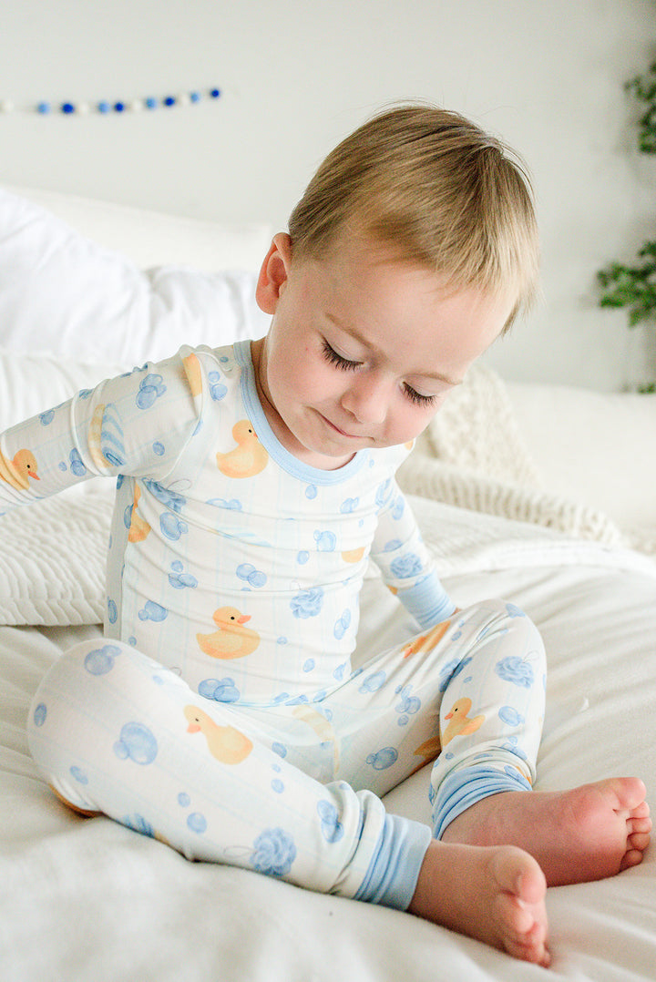 Boy sitting on bed looking down at blue Bubble Duckies two-piece bamboo pajamas showing full outfit