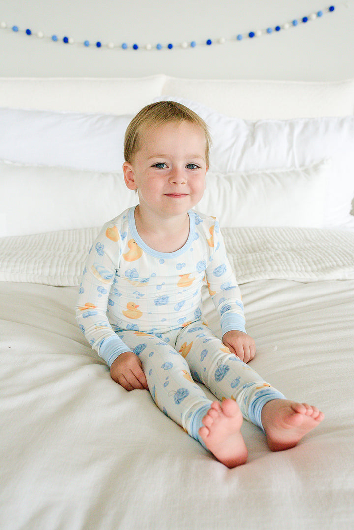 Toddler sitting on bed wearing blue Bubble Duckies two-piece bamboo pajamas with yellow duck print