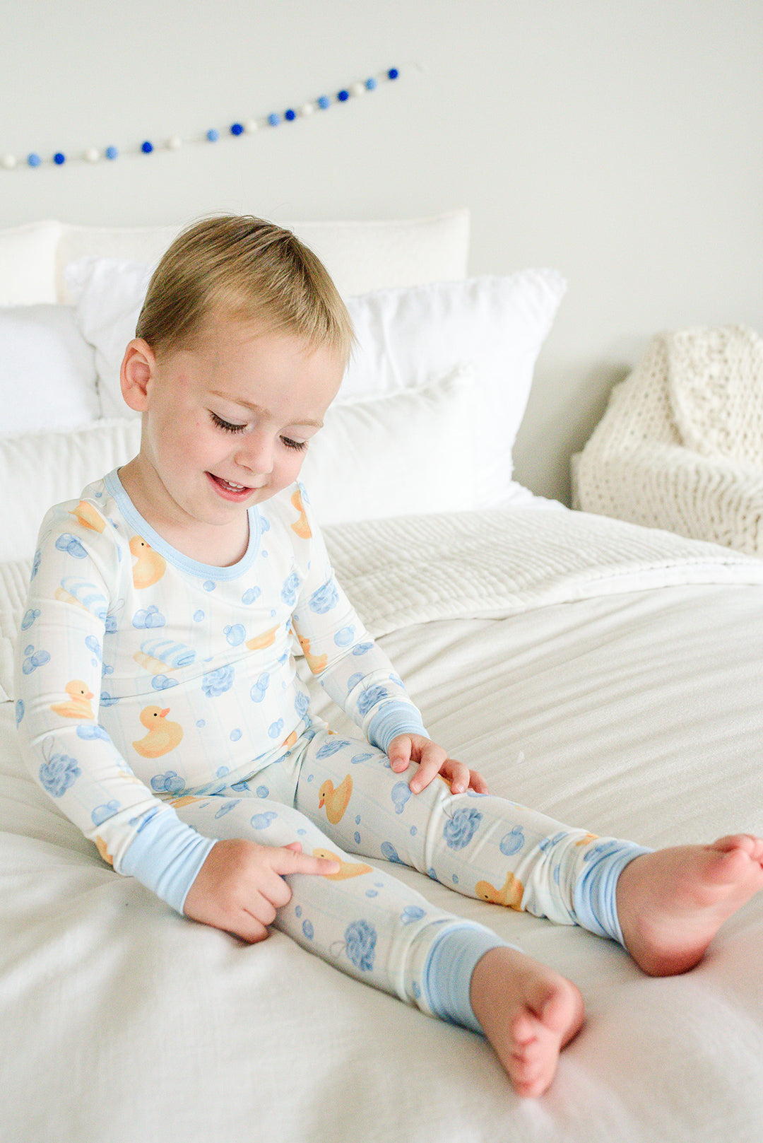 Young boy sitting on bed in blue Bubble Duckies two-piece bamboo pajamas looking at his feet