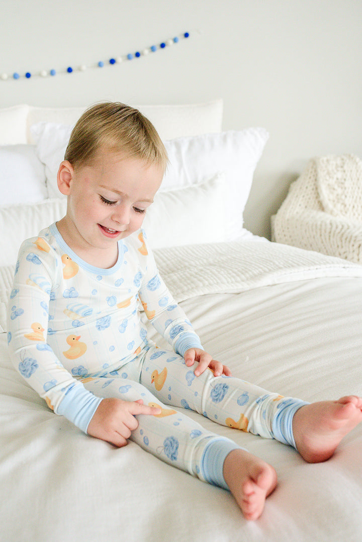 Young boy sitting on bed in blue Bubble Duckies two-piece bamboo pajamas looking at his feet