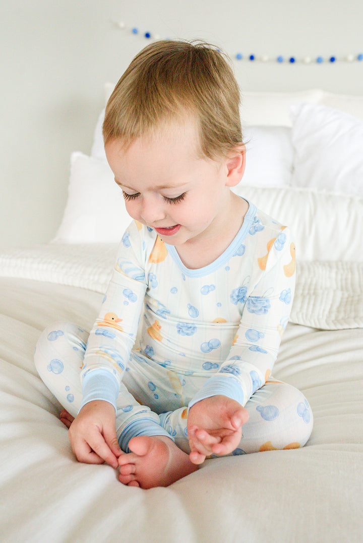 Toddler sitting cross-legged on bed in blue Bubble Duckies two-piece bamboo pajamas with duck print