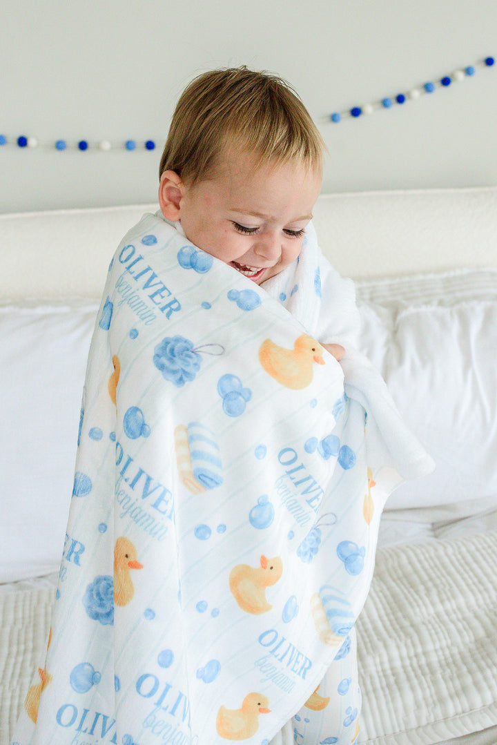 Young boy wrapped in blue Bubble Duckies personalized kids blanket with yellow rubber duck and blue bubble print featuring custom name