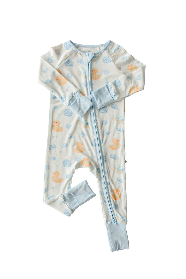 Baby boy wearing blue Bubble Duckies convertible zip romper with duck print sitting on couch with stuffed duck toy