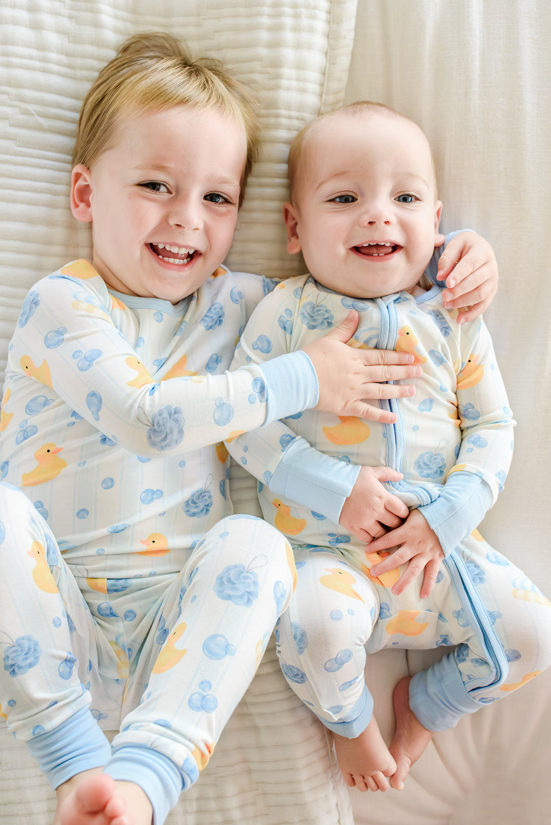 Two babies lying together wearing blue and pink Bubble Duckies zip romper pajamas showing matching colors