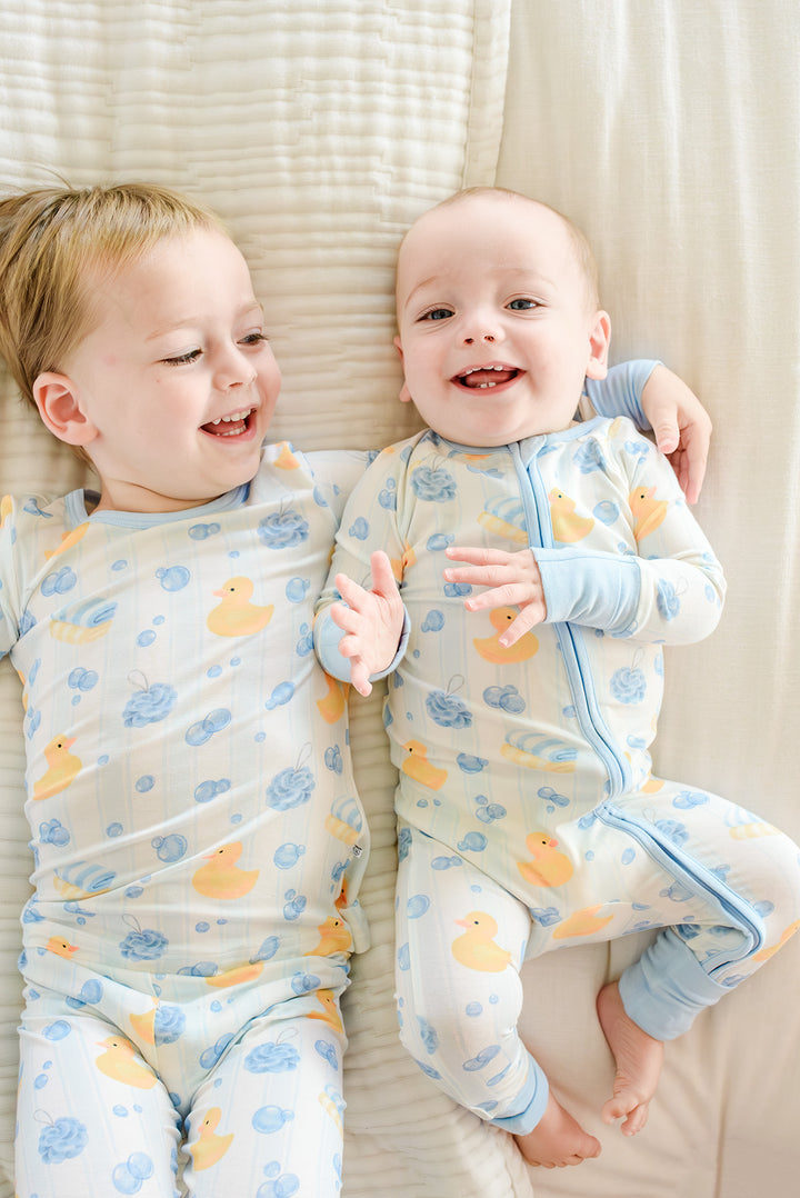 Two babies lying together in matching blue and pink Bubble Duckies bamboo zip romper pajamas
