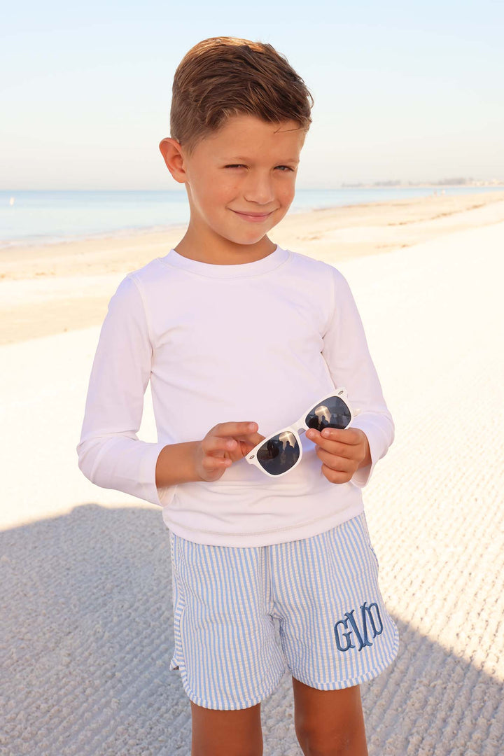 boy standing on the sand in monogrammed blue seersucker swim trunks with white long sleeve rash guard shirt 