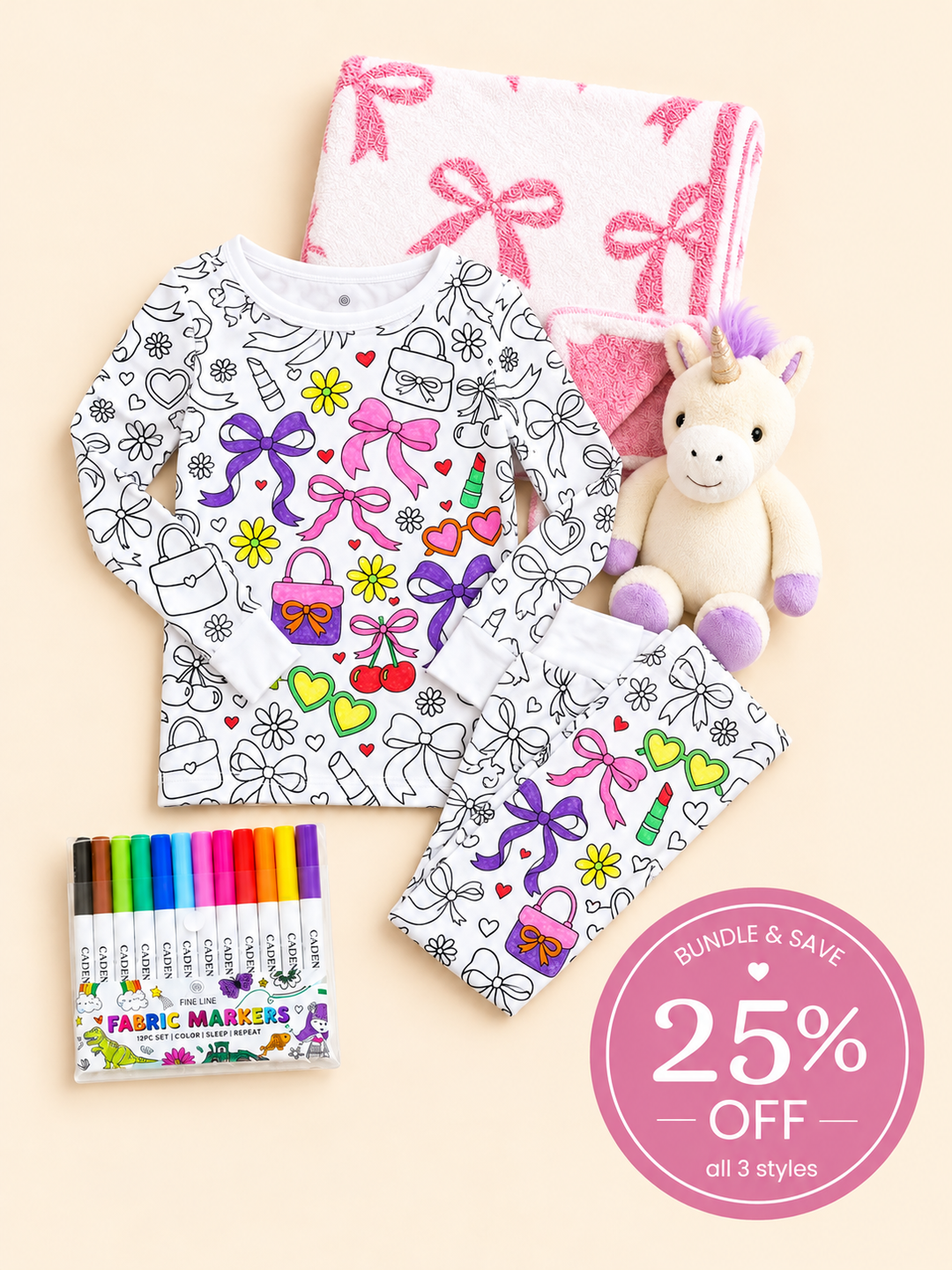 Flat lay of a Caden Lane color-in bow pajama set beside a folded pink bow blanket, paired with a plush unicorn and fabric markers.