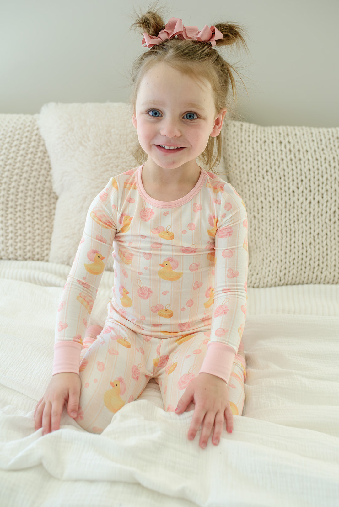 Young girl sitting on bed wearing pink Bubble Duckies two-piece bamboo pajama set with long sleeve top and pants
