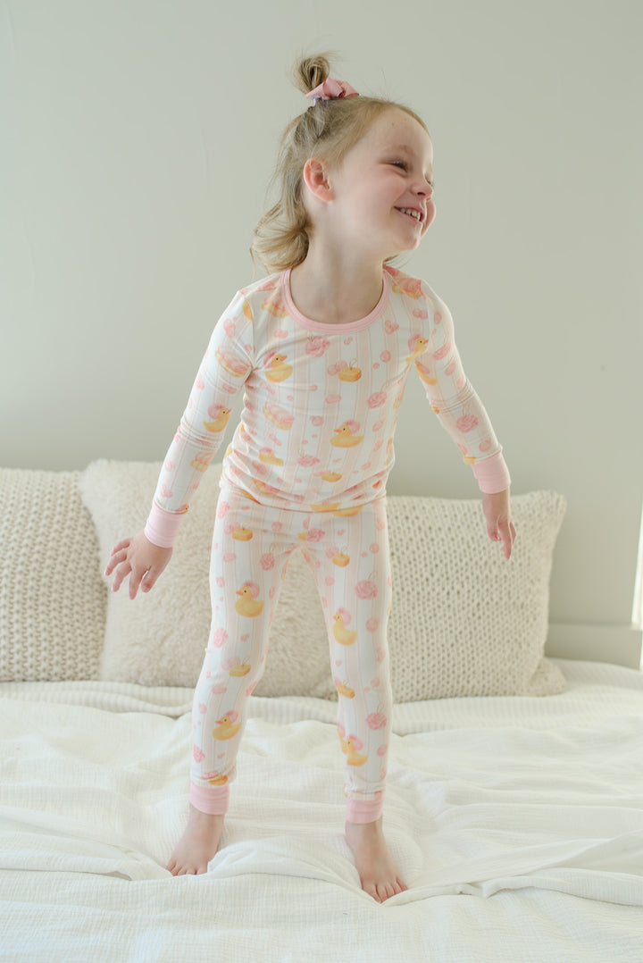 Young girl jumping on bed wearing pink Bubble Duckies two-piece bamboo pajama set with yellow duck print