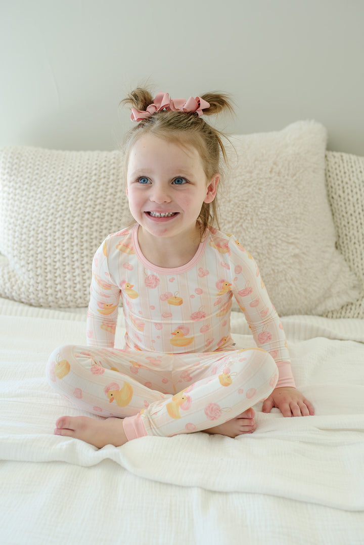 Girl sitting cross-legged on bed in pink Bubble Duckies two-piece bamboo pajamas smiling at camera