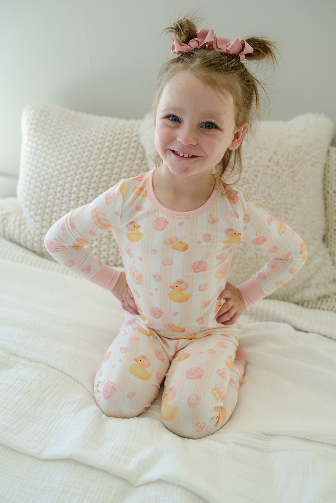 Smiling girl sitting on bed with hands on hips wearing pink Bubble Duckies two-piece bamboo pajamas set