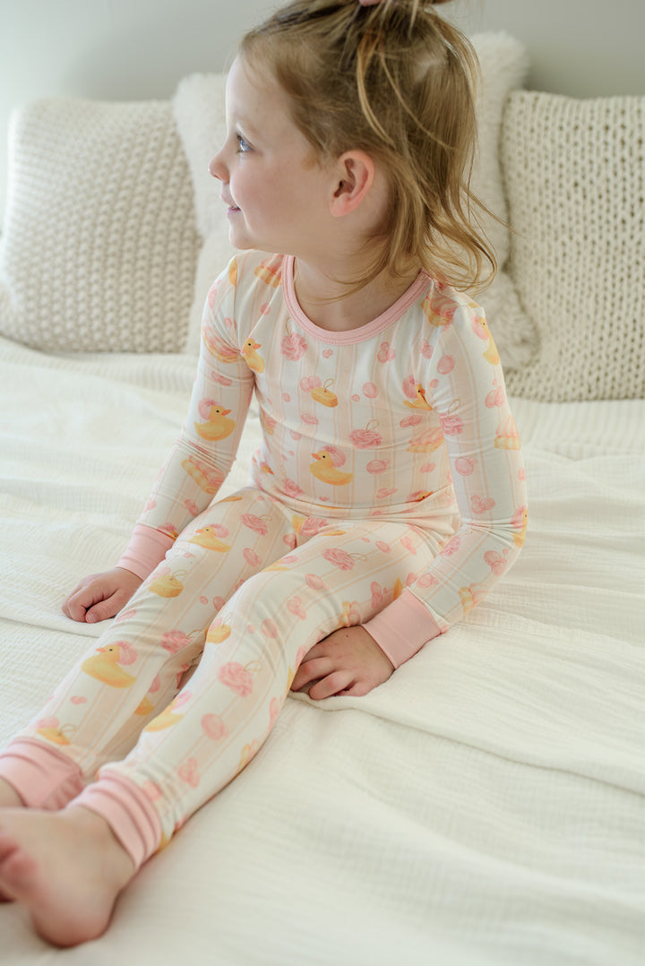 Young girl sitting on white bed in pink Bubble Duckies bamboo pajamas looking sideways, showing full set