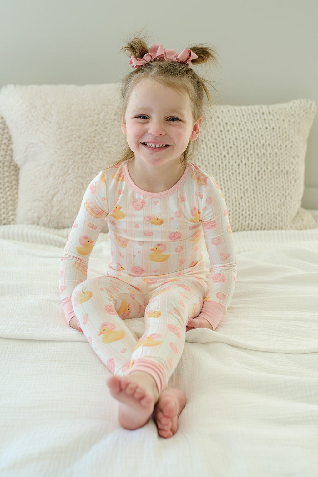 Smiling girl sitting on bed in pink Bubble Duckies two-piece bamboo pajamas with pigtails and pink hair ties