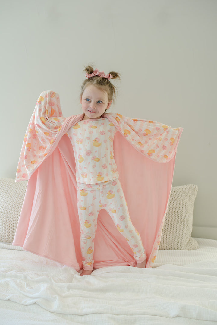 Young girl in matching pajamas holding wide open pink Bubble Duckies bamboo blanket showing printed and solid pink reverse sides