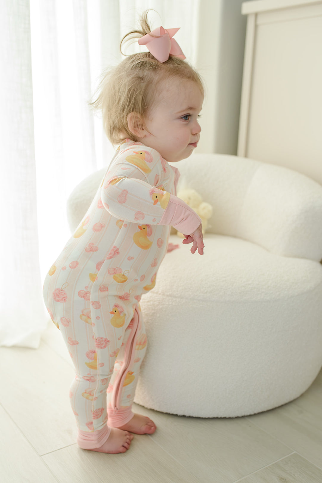 Baby girl with pink bow in pink Bubble Duckies zip romper sitting on matching rubber duck print bedding