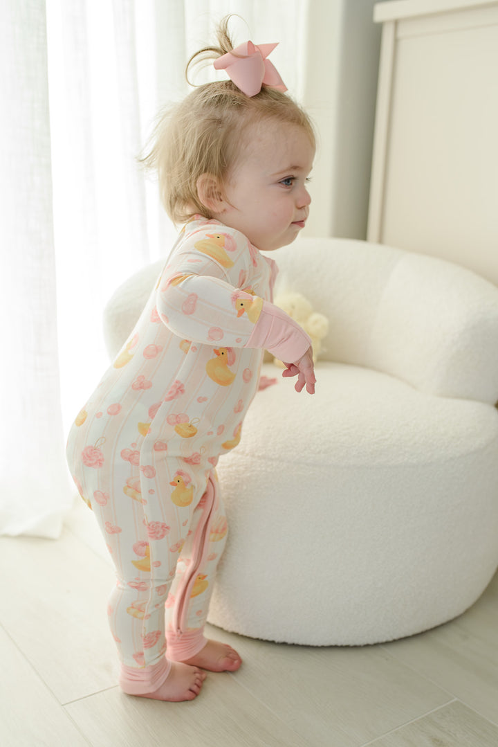 Baby girl with pink bow in pink Bubble Duckies zip romper sitting on matching rubber duck print bedding