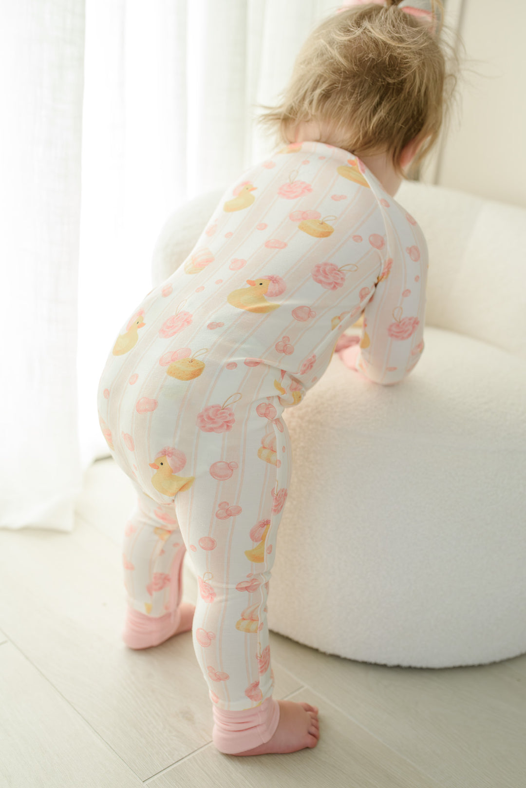 Adorable baby girl in pink Bubble Duckies zip romper on matching bedding with pink bow clapping